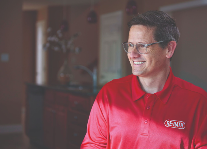 A smiling man wearing a red Re-Bath polo shirt stands in a kitchen. | Re-Bath