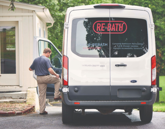 A Re-Bath installer steps into the driver’s seat of a large van with the company logo printed on the rear windshield. | Re-Bath