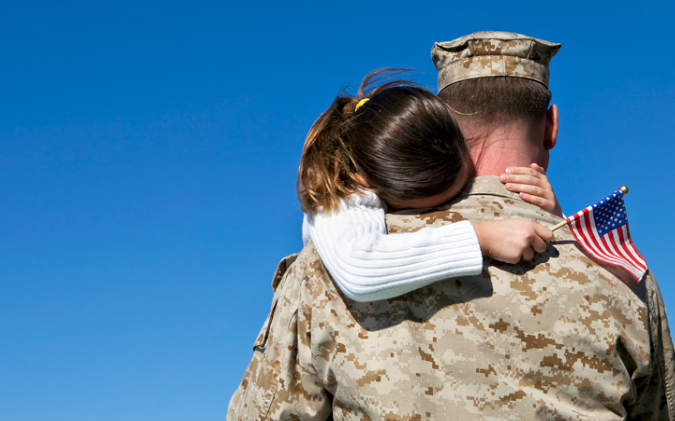 A military member in uniform embraces a young girl holding a small American flag. | Re-Bath Franchising