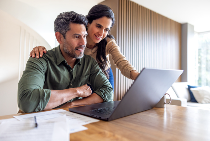 A couple sits at a wooden table in a modern home, reviewing documents together on a laptop. | Re-Bath