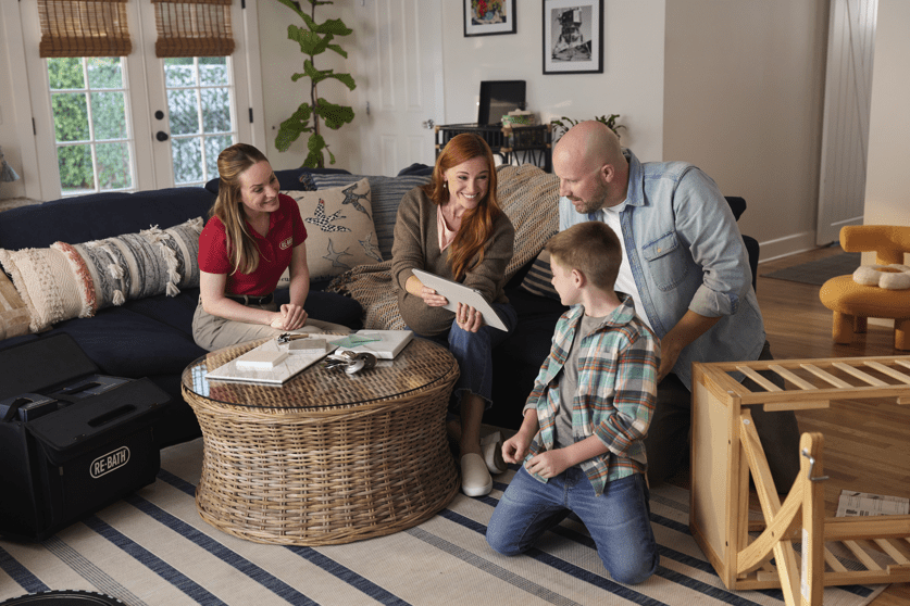 A consultant meets with a smiling family in their living room, showing renovation plans on a tablet. | Re-Bath